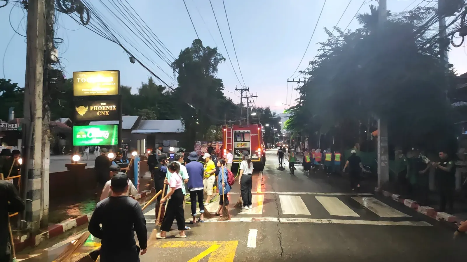 Volunteer cleanup crew sweeping a flooded street at dusk with a fire truck pressure-washing mud off the road